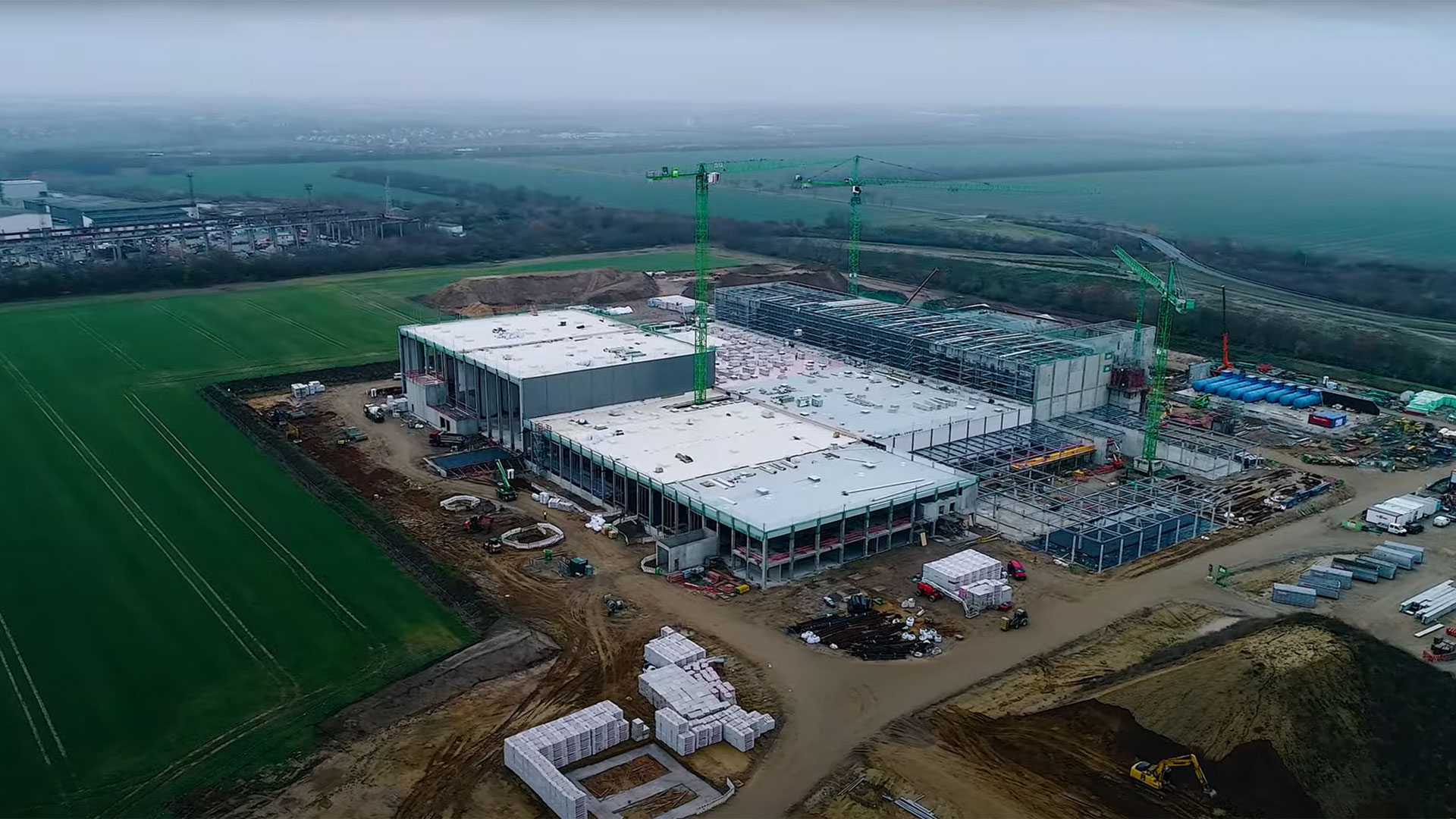 Bau einer modernen Industrieanlage mit Kränen und Baustellenfahrzeugen in einer ländlichen Umgebung.