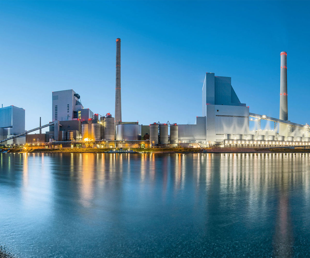 Industrieanlage mit hohen Schornsteinen und großen Lagertanks, reflektiert im Wasser bei Dämmerung.