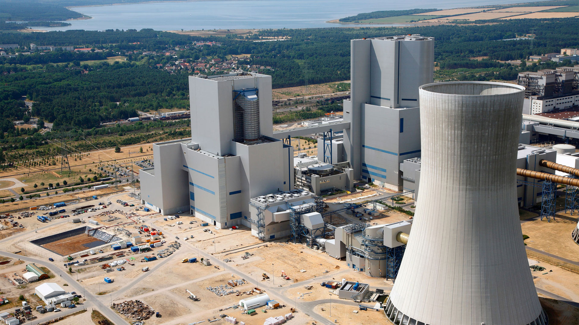 Luftaufnahme eines Kraftwerks mit Kühlturm und mehreren großen Gebäuden in einer industriellen Umgebung.